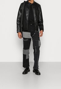 Hombre con chaqueta de cuero negra, camisa negra, jeans de parche negro con parches grises y estampados, zapatos negros y llevando una bolsa de hombro negra.