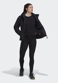 Black padded jacket with hood, worn open over a black top, paired with black leggings and athletic shoes, featuring a sleek, streamlined design.