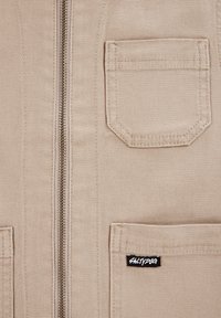 Beige jacka med dragkedja framtill, två bröstfickor och en etikett längst ner till höger. Tyget är strukturerat och hållbart.