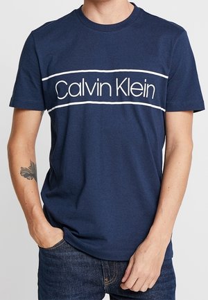 T-shirt bleu marine à manches courtes avec logo blanc « Calvin Klein » et rayures horizontales porté par une personne avec un tatouage sur l'avant-bras gauche.