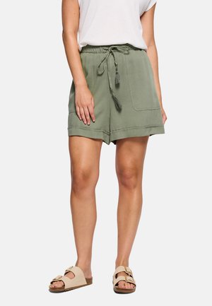 Mujer con pantalones cortos de cordón verde oliva con bolsillos delanteros y sandalias beige con hebilla, de pie contra un fondo liso.