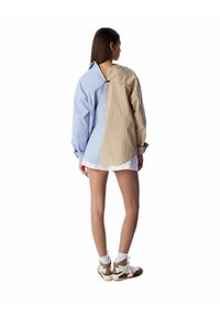 Camicia oversized bicolore con pannelli a righe blu e beige. Il retro presenta un taglio aperto e le maniche sono a polsino.