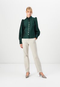 Woman wearing a dark green textured blouse with gold buttons and puff sleeves, light beige pants, and pointed grey heels standing against white background.