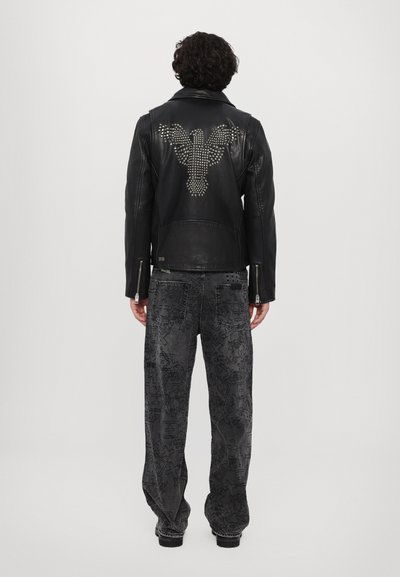Svart läderjacka med nitar och örn-design på ryggen, kombinerad med löst sittande mörka jeans med intrikata mönster.