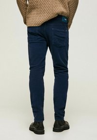 Man som bär marinblå smal passform corduroy byxor, svarta stövlar och en brun kabelstickad tröja, stående framför en enkel bakgrund.