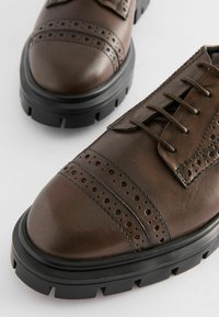 Hnědé kožené boty s wingtip designem, kulatými perforacemi, robustní černou gumovou podrážkou a šněrováním pro bezpečné uchycení.