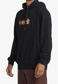 Hombre con sudadera negra con capucha y diseño floral naranja en el pecho y el bolsillo delantero, combinado con pantalones marrones, sobre fondo blanco.