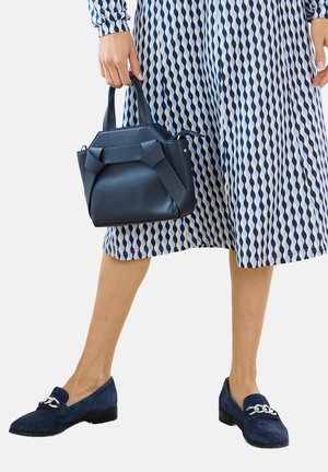 Borsa a mano blu scuro con texture e design strutturato, abbinata a un vestito blu e bianco a fantasia e mocassini blu.