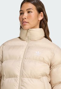 Beige puffer jacket with a high collar, featuring a full zipper and subtle horizontal stitching. Notable white Adidas logo on the chest.