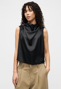 Μαύρο σατέν αμάνικο τοπ με ντραπαρισμένο ντεκολτέ; το ύφασμα έχει απαλή υφή. Συνδυάζεται με μπεζ ψηλόμεσα παντελόνια.