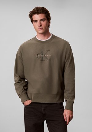 Mann trägt olivgrünes Calvin Klein Sweatshirt und schwarze Hose, steht mit einer Hand in der Tasche vor einfarbigem grauem Hintergrund.