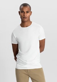T-shirt bianco a maniche corte realizzato in tessuto morbido, con scollo rotondo e silhouette aderente. Abbinato a pantaloni beige.