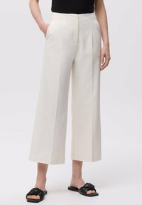 Pantaloni a gamba larga in tessuto crema, con vita alta, tasche laterali e una texture liscia. Sandali neri con dettaglio a nodo.