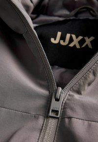 Podrobnosti sive jakne, ki vključujejo zadrgo, gladko teksturo in vidno oznako "JJXX" na črni notranjosti. Potegalo zadrge ima pravokotno obliko.