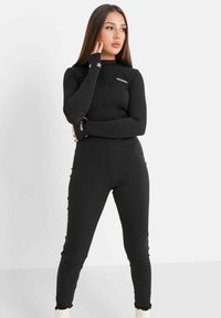 Schwarzer, langärmeliger Jumpsuit mit hohem Kragen und tailliertem Design. Mit geripptem Textur und kontrastierender Markenkennung an den Bündchen.