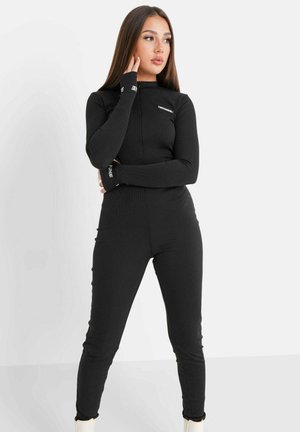 Zwarte jumpsuit met lange mouwen, hoge hals en een getailleerd ontwerp. Kenmerkt zich door ribbeltextuur en contrasterende branding op de manchetten.