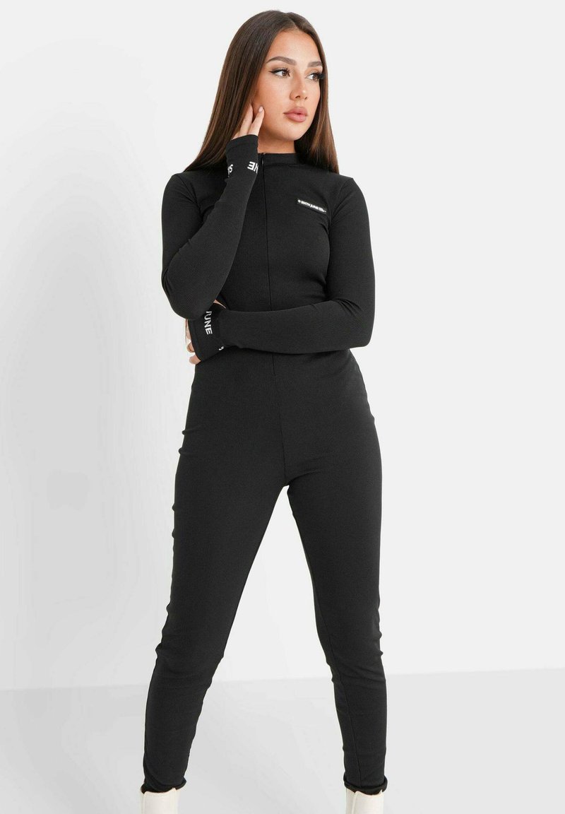 Schwarzer, langärmeliger Jumpsuit mit hohem Kragen und tailliertem Design. Mit geripptem Textur und kontrastierender Markenkennung an den Bündchen.