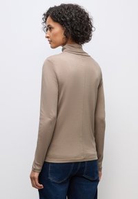 Beige Langarmshirt mit Stehkragen, glatter Textur, taillierter Passform und einer Naht, die mittig am Rücken verläuft. Kombiniert mit dunklen Jeans.