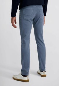 Man die slim-fit blauwe jeans en witte sneakers draagt, staand tegen een effen lichte achtergrond, gezien vanaf de achterkant.