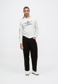 Maglietta a maniche lunghe grigio chiaro con design cartoon, abbinata a pantaloni in corduroy marrone scuro. Sneakers bianche con dettagli grigi.