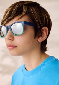 Blauwe zonnebril met een transparant ombre montuur en reflecterende lenzen, gedragen door een kind met kort bruin haar, in een lichtblauw shirt.