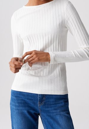 Personne portant un haut blanc à manches longues côtelé et un jean bleu, les mains ajustant la manche au poignet.