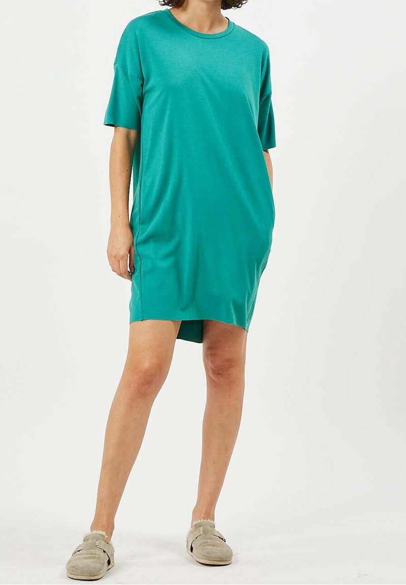 Femme portant une robe ample turquoise à manches courtes avec poches et des chaussures beige à enfiler, debout devant un fond clair uni.