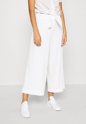 Femme portant un pantalon blanc large et court, une chemise blanche nouée, et des baskets blanches avec de légers accents jaunes, debout devant un fond uni.