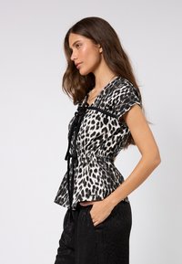 Svart-hvitt leopardmønstret topp med korte ermer, peplumstil og svart bånd som knytes i halsringen, brukt med mørke bukser.