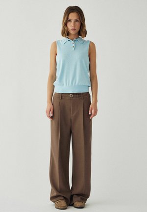 Kvinde står mod ensfarvet baggrund iført lyseblå ærmeløs polo top, brune viddebukser med bælte og brune mokkasinsko.