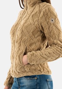 Giacca in pile color beige con un motivo a diamante testurizzato, colletto alto e chiusura con zip. Include tasche laterali e polsini a coste. Visibili jeans in denim.