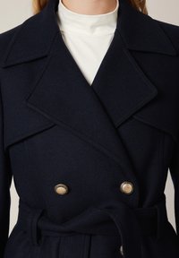 Granatowy wełniany płaszcz z dużymi, klapami, podwójnym zapięciem, złotymi guzikami i przewiązywanym paskiem w talii. Gładka faktura, dopasowany krój.