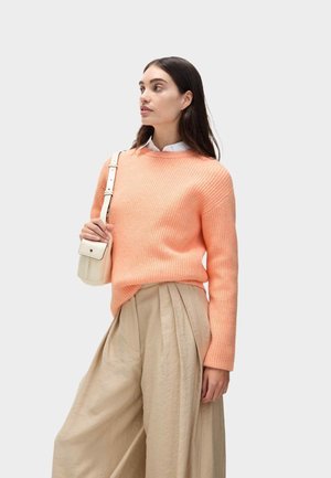 Vrouw met lang donker haar, gekleed in een perzikkleurige trui, beige broek met wijde pijpen, wit shirt en een kleine witte schoudertas dragend.