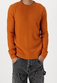 Pomarańczowy sweter z dzianiny z okrągłym dekoltem, prążkowanymi mankietami i krawędzią, noszony na luźnych szarych dżinsach z widocznymi kieszeniami i wyblakłą fakturą.