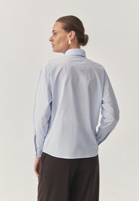Light blue striped button-up shirt with long sleeves, featuring a collar and a curved hem, paired with dark trousers. Gold earrings visible.