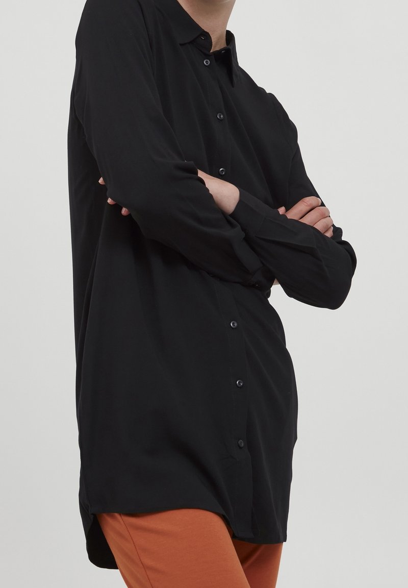 Chemise noire boutonnée en tissu lisse, avec col, manches longues et ourlet arrondi, associée à un pantalon orange.