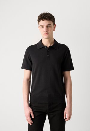 Jeune homme aux cheveux bouclés portant un polo noir à manches courtes et un pantalon noir, debout devant un fond blanc uni.