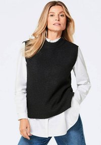 Zwarte gebreide mouwloze trui over een witte overhemd, met een hoge halslijn en geribbelde structuur, gecombineerd met blauwe jeans.