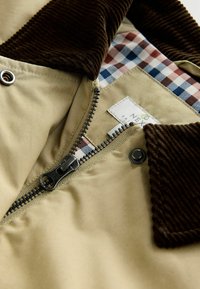 Giacca tan con colletto in corduroy marrone, chiusura con zip e fodera a scacchi blu, rossa e bianca, con un'etichetta all'interno.