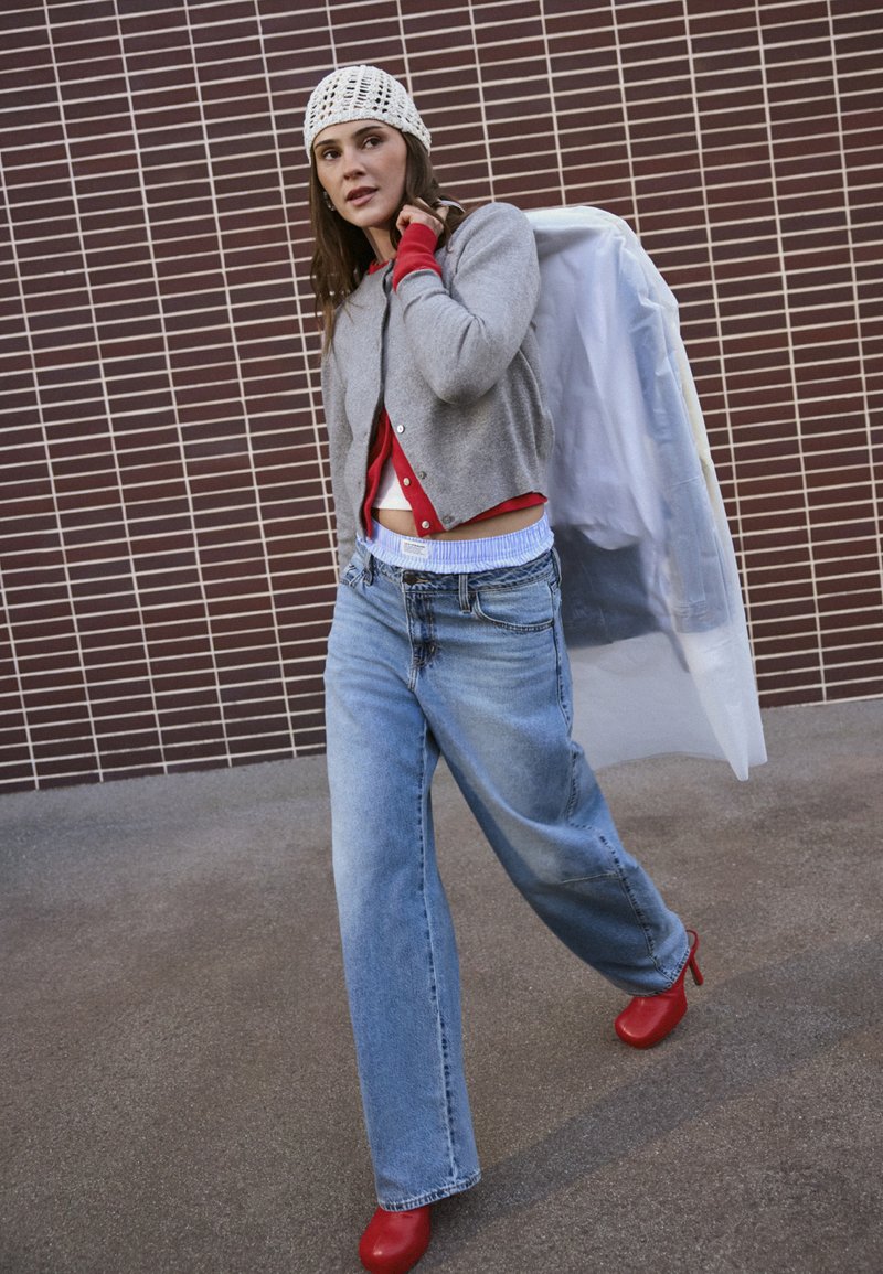 Frau geht an einer gefliesten Wand vorbei, trägt eine graue Strickjacke, Jeans mit weitem Bein, rote Schuhe, eine weiße Strickmütze und trägt eine Kleiderhülle über der Schulter.