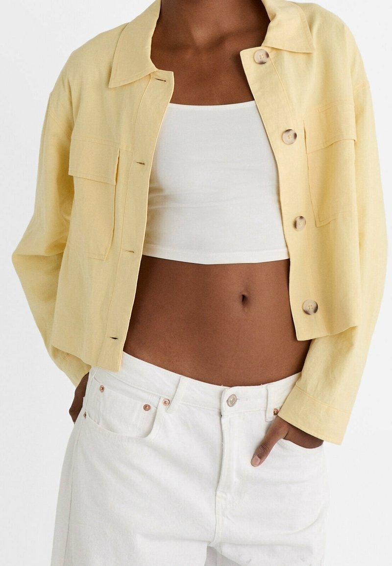 Veste chemise en lin jaune avec col, boutons et poches avant, portée avec un crop top blanc et un jean blanc. Texture lisse.