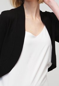 Černý krátký cardigan s hladkou pletenou texturou, s otevřeným předním dílem a krátkými rukávy, nošený přes bílý top s výstřihem do V.