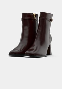 Para ciemno-brązowych skórzanych botków za kostkę z szerokimi obcasami, bocznymi zamkami błyskawicznymi oraz ozdobnymi paskami z małymi złotymi klamrami.