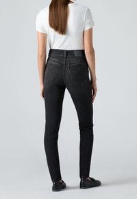 Femme aux cheveux longs et raides portant une chemise blanche à manches courtes, un jean skinny noir taille haute et des mocassins noirs, debout de dos.