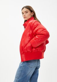 Rød pufferjakke med høj krave, blank overflade og ribbede mansjetter. Har lynlås og sidelommer, båret over blå denimjeans.