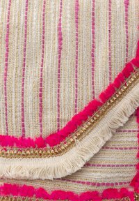 A textil párnahuzat krémszínű alapja arany cérnával, rózsaszín kiegészítőkkel és texturált bojt részletekkel rendelkezik a széleken.