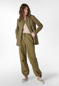 Giacca verde khaki con texture e bordi sfilacciati sopra un top beige a coste e pantaloni cargo larghi, abbinati a sneakers beige con lacci.