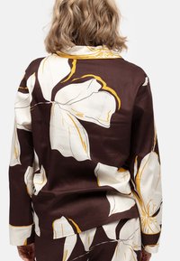 Camisa floral marrón con grandes patrones de flores blancas y amarillas, con mangas largas, cuello y un corte relajado. Fabricada en tela suave.