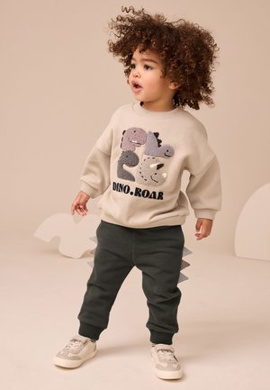 Toddler med krøllet hår står iført beige dinosaur-sweatshirt, mørke bukser med hale pigge og hvide sneakers mod ensfarvet baggrund.