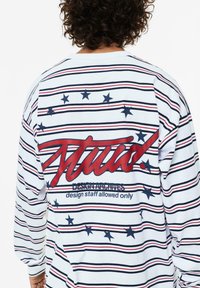 Langärmliges Hemd in Weiß mit roten und marineblauen Streifen, mit einem großen Grafikdesign aus roten und blauen Sternen auf dem Rücken, aus Baumwollstoff gefertigt.